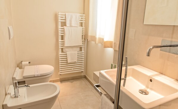 Un baño moderno con un lavamanos, inodoro y bañera blancos. Hay un perchero con varias toallas, un box de ducha con paredes de vidrio y un espejo sobre el lavamanos.
