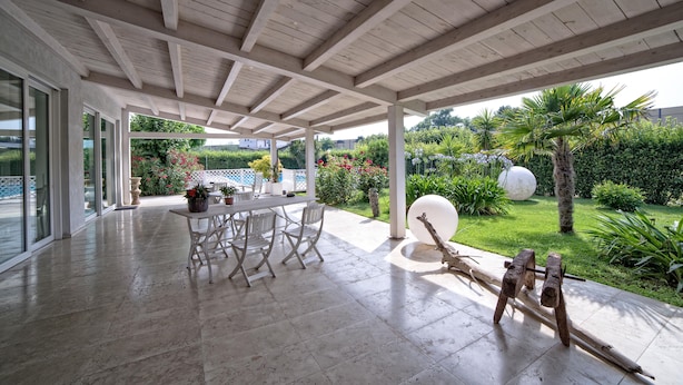A covered patio with a table and chairs, a sculpture, and a view of a garden with a palm tree and a large white spherical object. - Hotel Radice (Civitanova Marche)