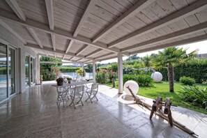 Un patio techado con una mesa y sillas, una escultura y vistas de un jardín con una palmera y un objeto blanco esférico grande.