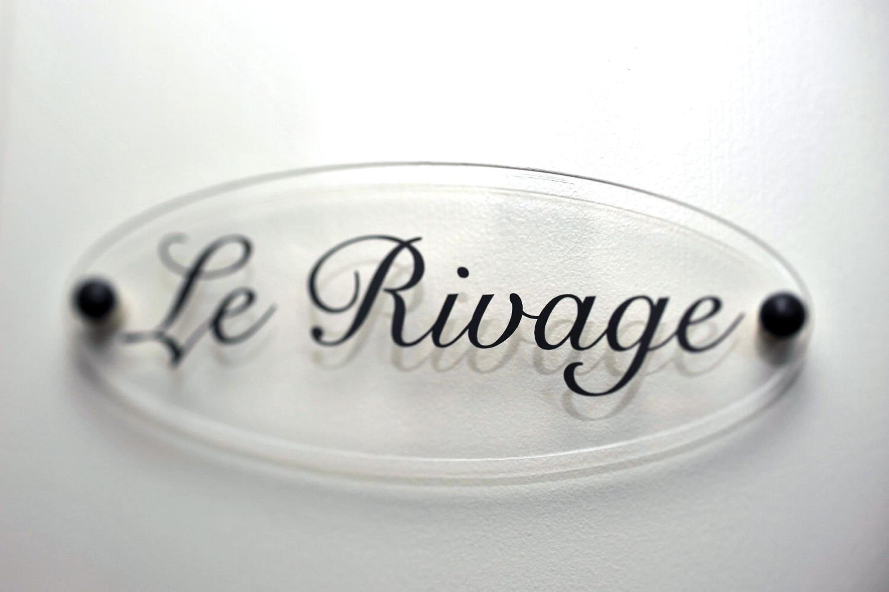 Een helder, ovaal bord met de tekst 'Le Rivage' in sierletters, bevestigd aan een witte muur met twee zwarte pennen.