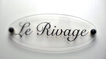 Een helder, ovaal bord met de tekst 'Le Rivage' in sierletters, bevestigd aan een witte muur met twee zwarte pennen.