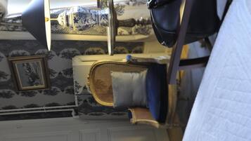 A room with patterned wallpaper, a bed with a blue headboard, a bedside table with a lamp, and a framed picture on the wall.