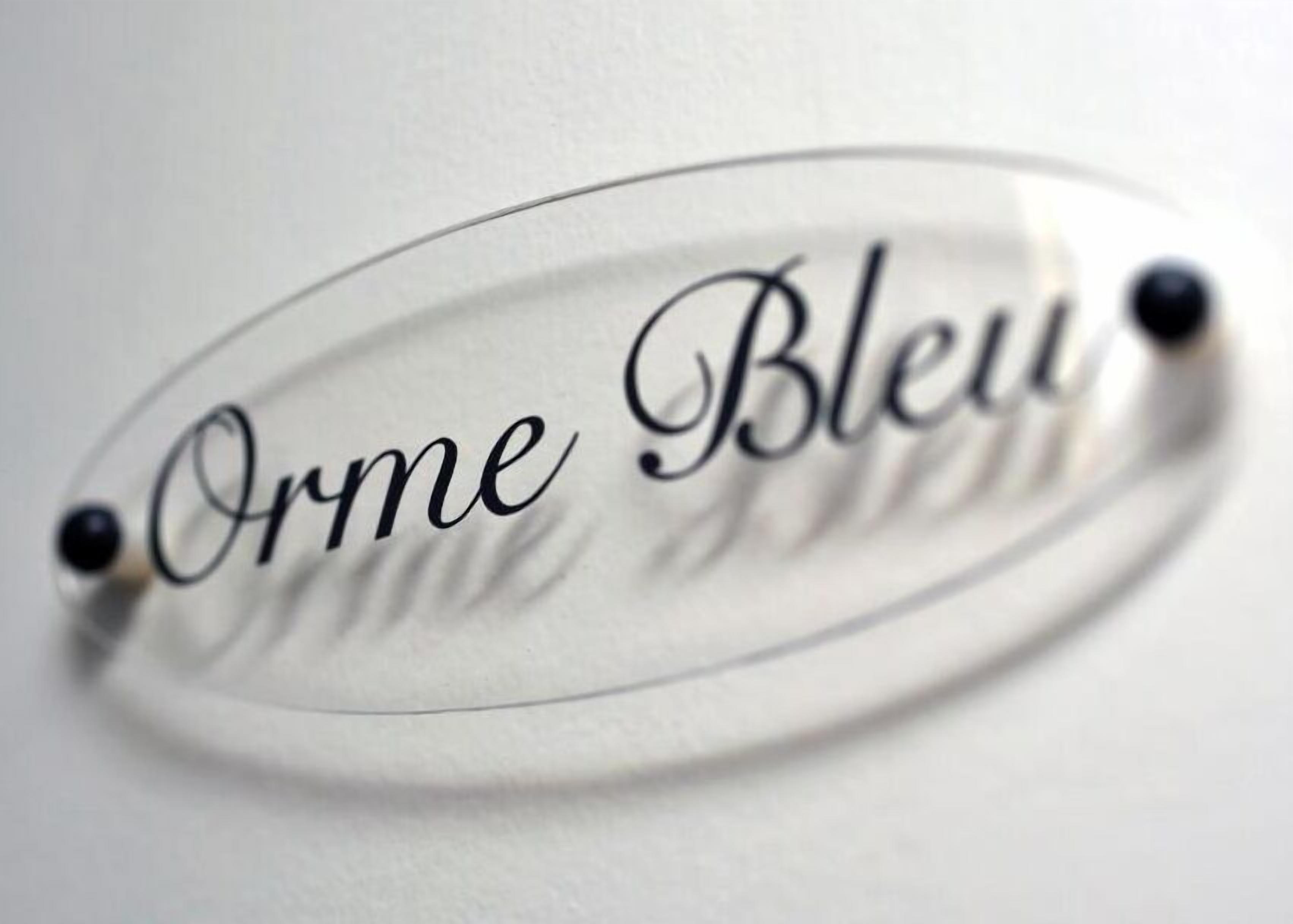 Een helder, ovaal bord met de tekst 'Orme Bleu' en aan weerszijden twee zwarte puntjes.