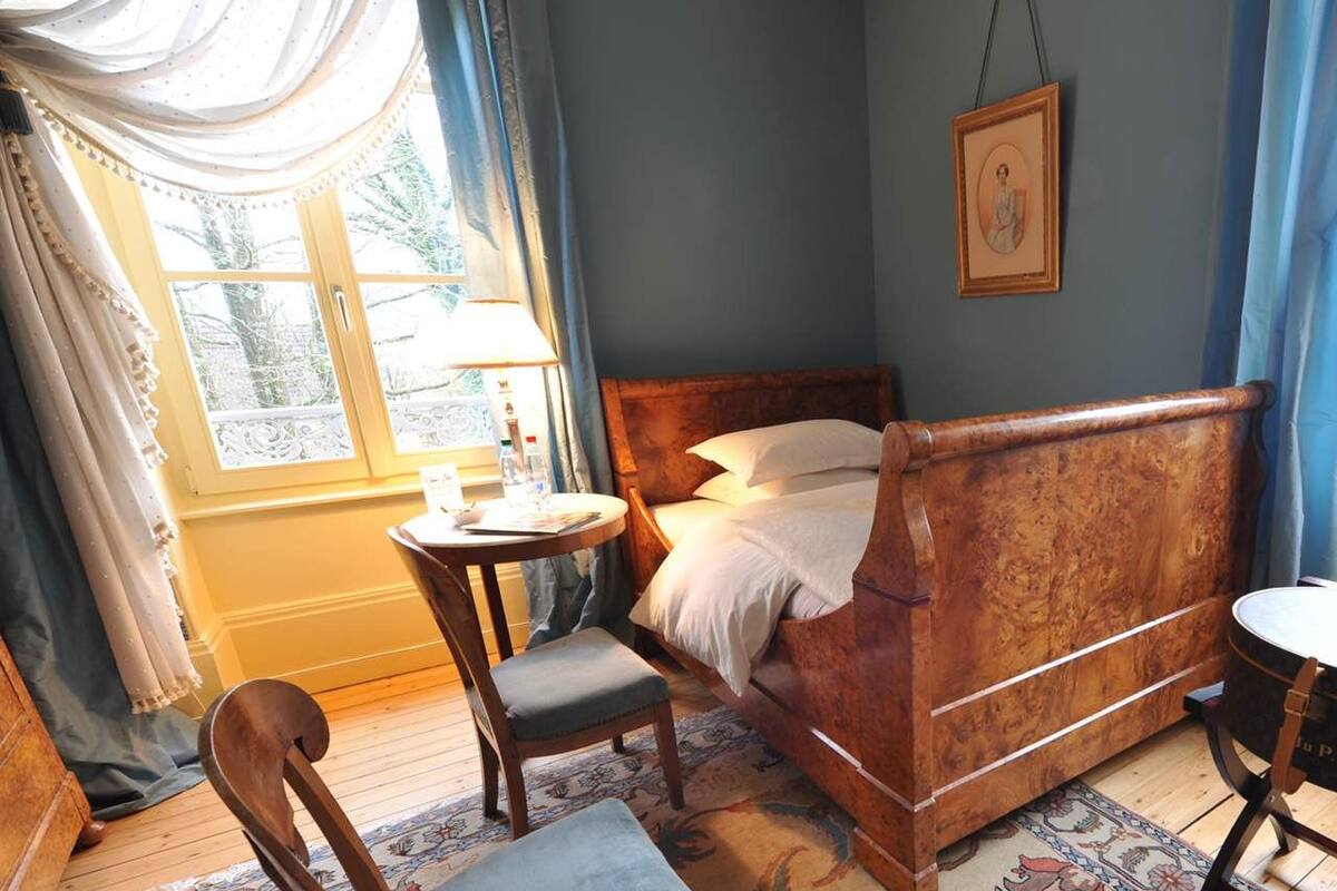 A traditional bedroom with a wooden four-poster bed, a round table with a lamp, and a chair.
