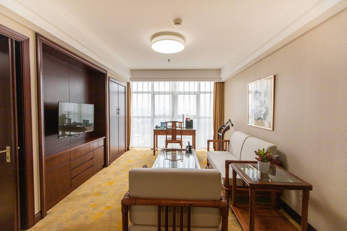 Une chambre d’hôtel avec un bureau en bois, un canapé, une télévision et une grande fenêtre avec des rideaux.