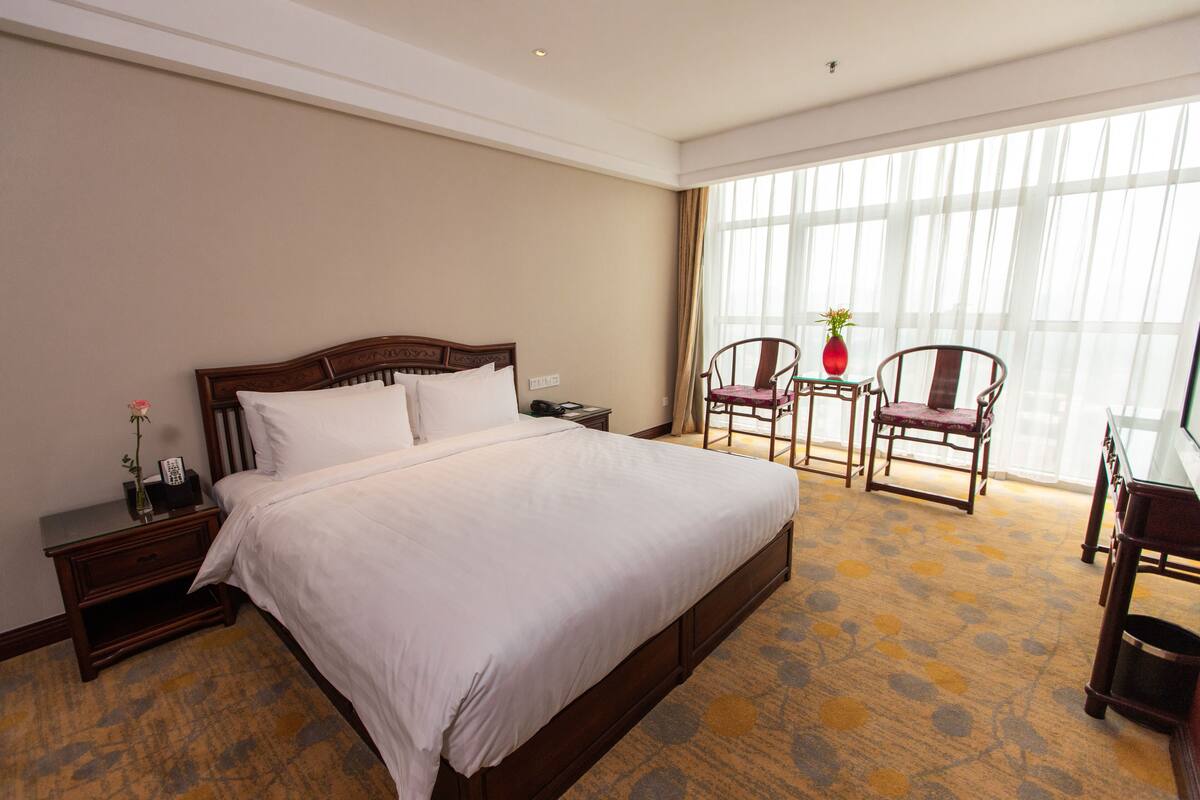 Une chambre d’hôtel avec un grand lit, une table de chevet, un bureau avec une chaise et une grande fenêtre avec des rideaux.