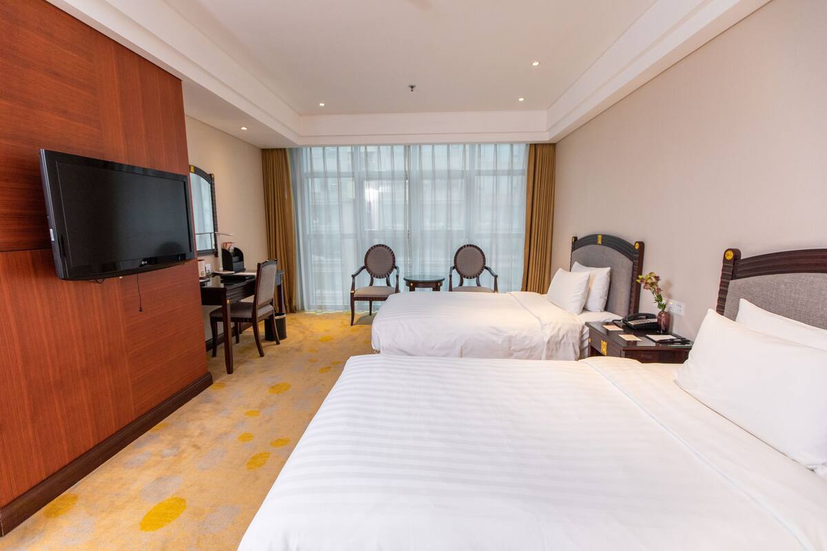 Une chambre d’hôtel avec deux lits, une télévision, un bureau avec une chaise et une grande fenêtre avec des rideaux.