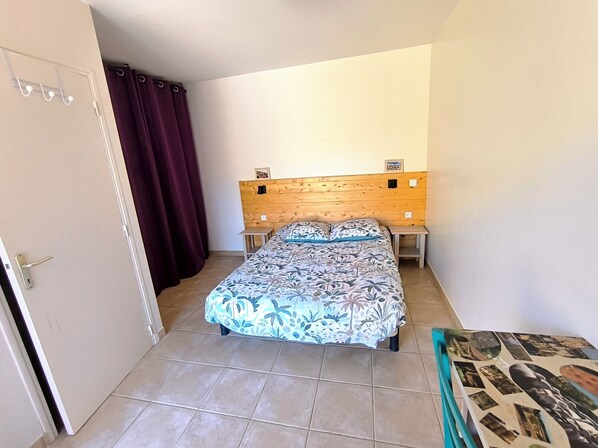 Un dormitorio con cama, cabecera de madera, mesita de noche pequeña, una puerta con picaporte y una cortina.