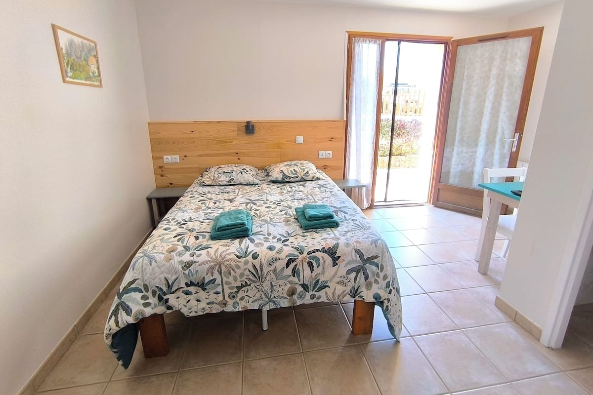 A bedroom with a bed, a nightstand, a chair, a window with curtains, and a door leading to an outdoor area.