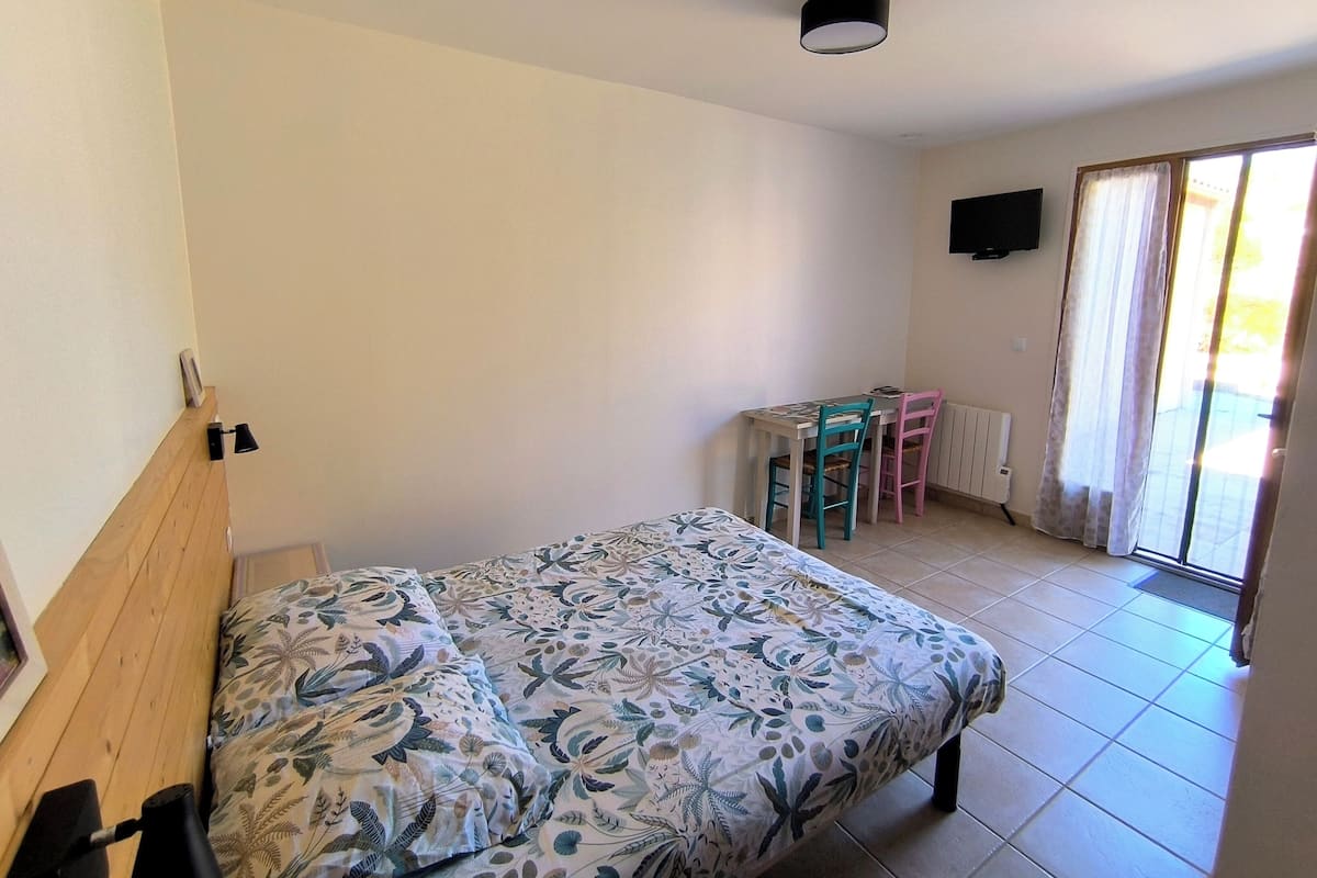 A bedroom with a bed, a desk, chairs, a radiator, and a TV on the wall.