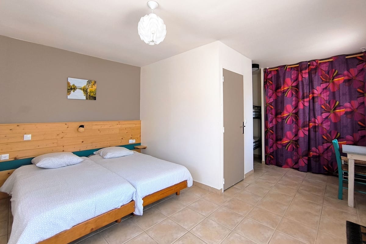 A bedroom with a wooden bed, a white ceiling light, a door leading to a bathroom, and a bathroom with a purple floral curtain.