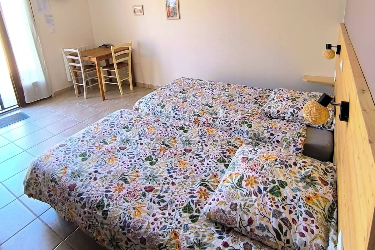 A bedroom with a floral bedspread, a wooden headboard, a desk with a chair, a television on the wall, and a ceiling light fixture.