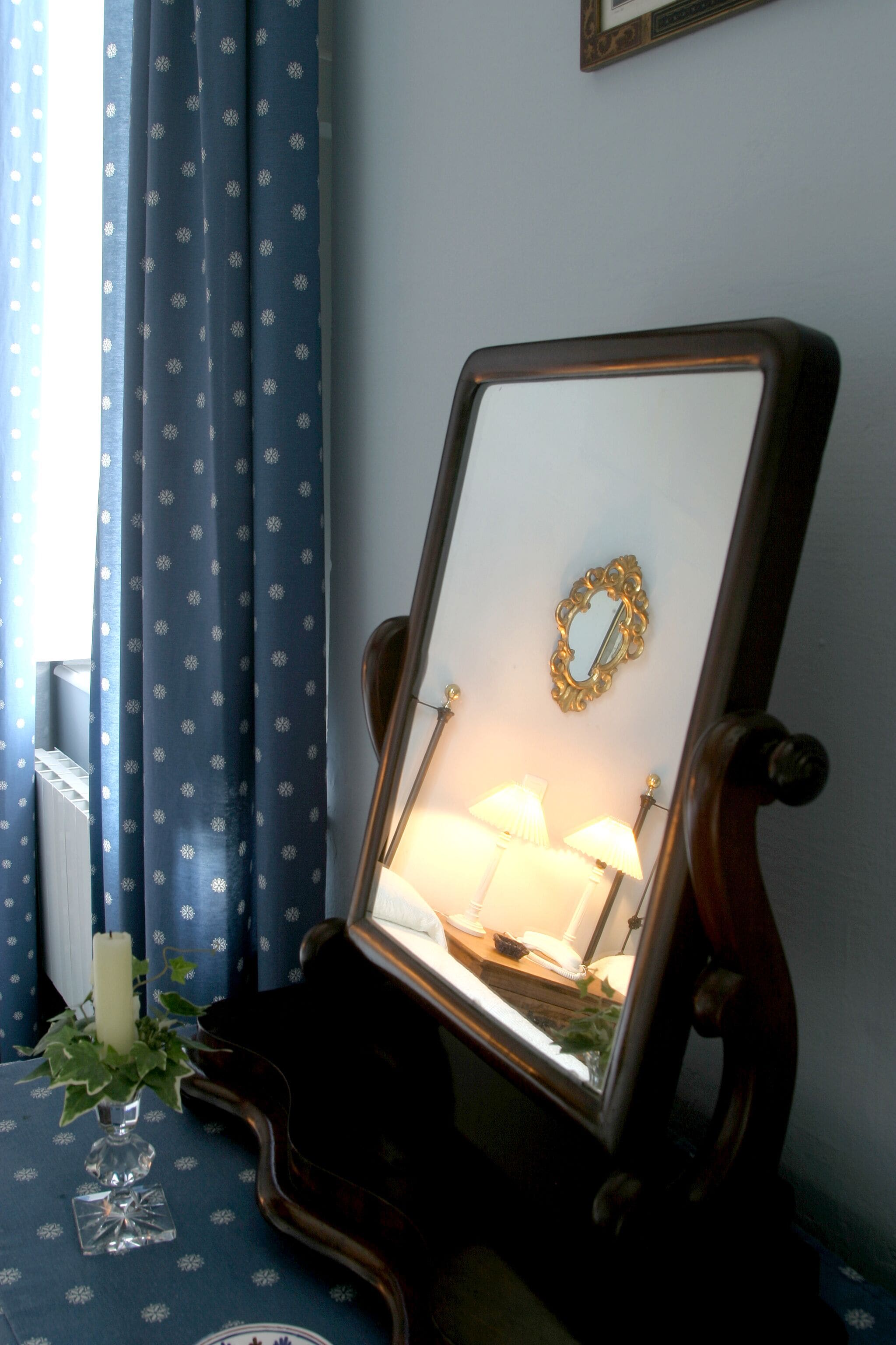 A mirror with a gold frame on a wooden stand, a white candle, and a vase with green foliage on a blue tablecloth.