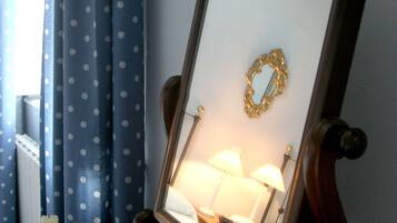 A mirror with a gold frame on a wooden stand, a white candle, and a vase with green foliage on a blue tablecloth.