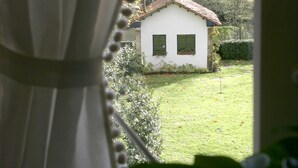 Vista a través de una ventana con cortina, mostrando una casa blanca con techo rojo, rodeada de vegetación.