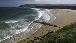 Un paisaje costero con una playa arenosa, un litoral rocoso y colinas cubiertas de hierba al fondo.