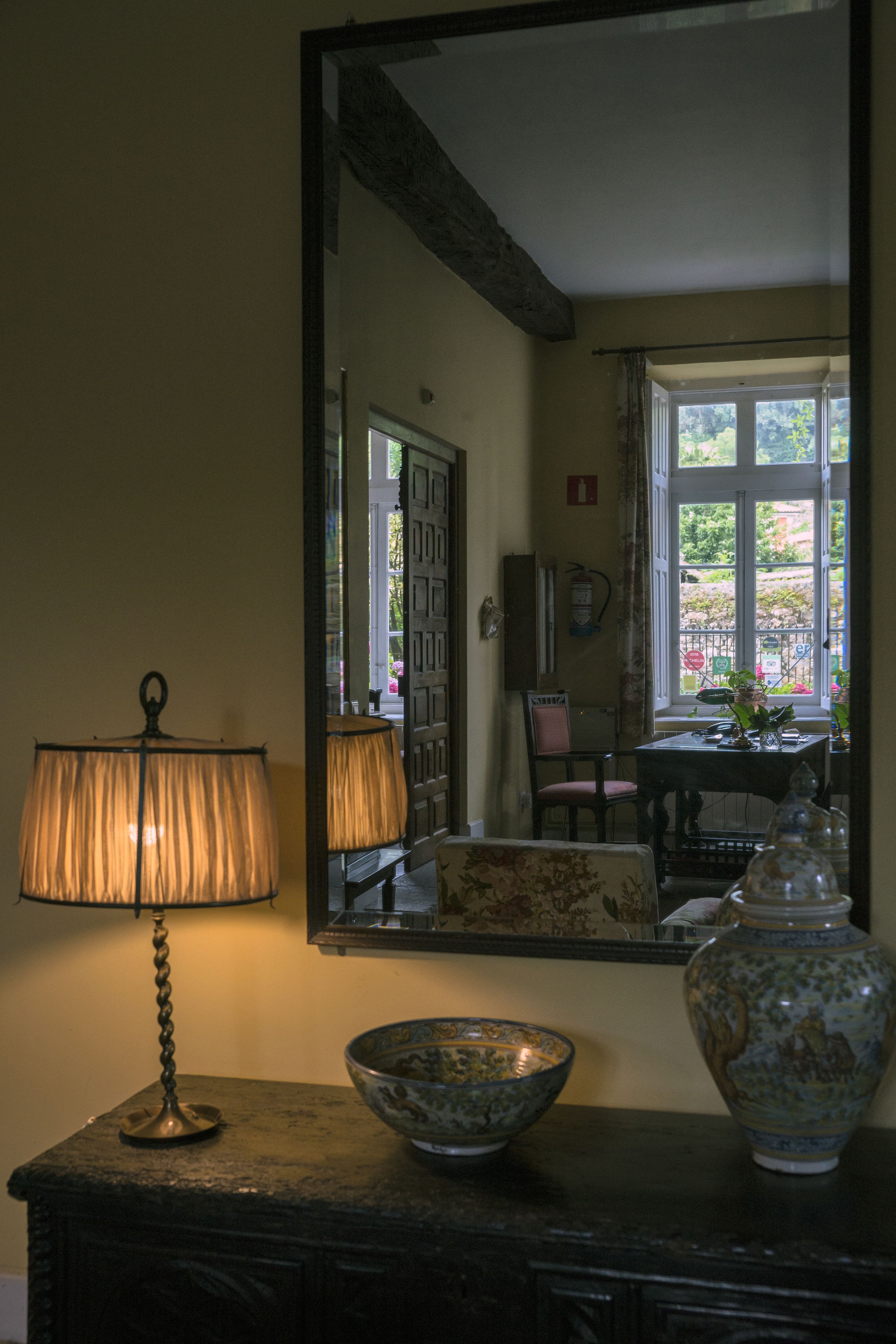 A room with a mirror reflecting a dining area, a lamp, a bowl, and a vase on a dresser.