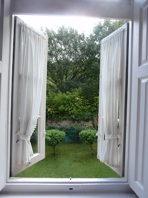 Janela aberta com cortinas brancas, mostrando vista de um gramado verde e árvores.