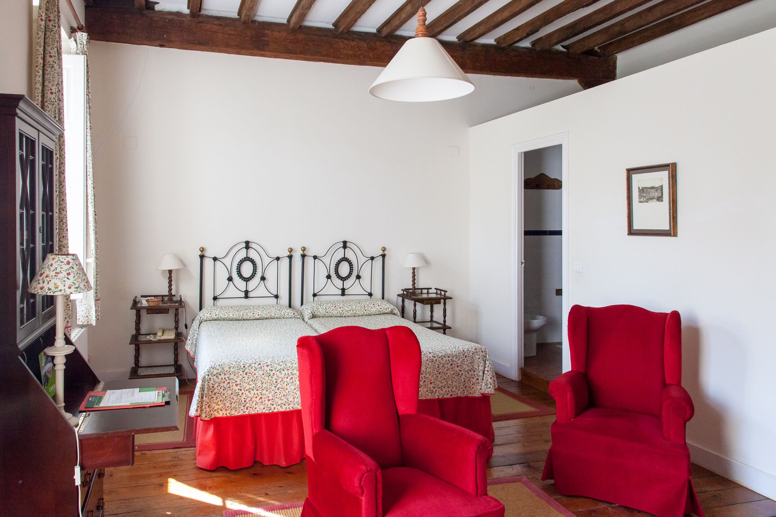 A bedroom with a wooden ceiling, a bed with a floral bedspread, two red chairs, a bedside table with a lamp, and a framed picture on the wall.