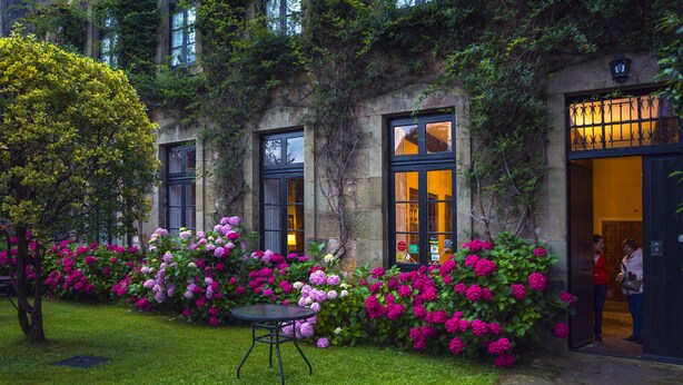 Un edificio con ventanas amplias, un jardín con flores rosadas y una pequeña mesa.