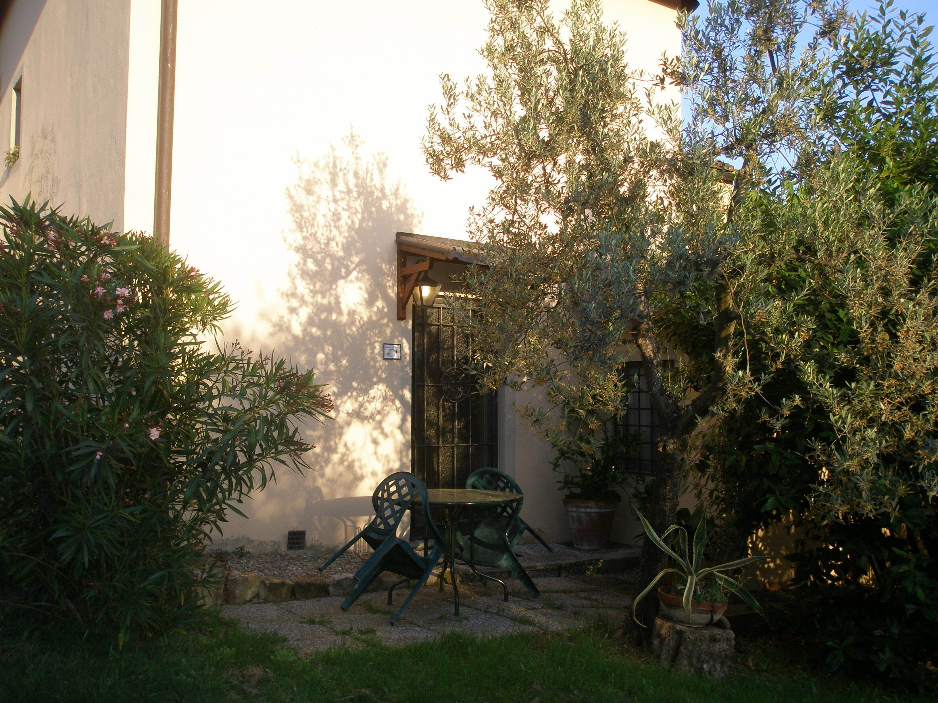 A courtyard with a table and chairs, surrounded by greenery and a building with a door and window.