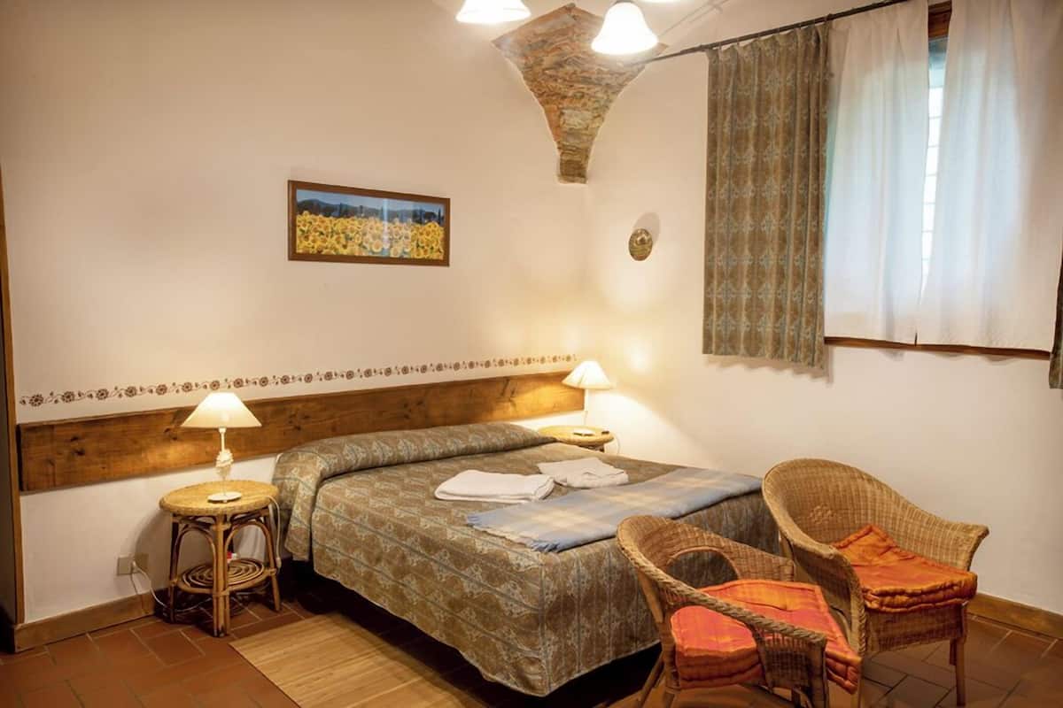 A bedroom with a bed, two wicker chairs, a small round table, a lamp, and a painting on the wall.