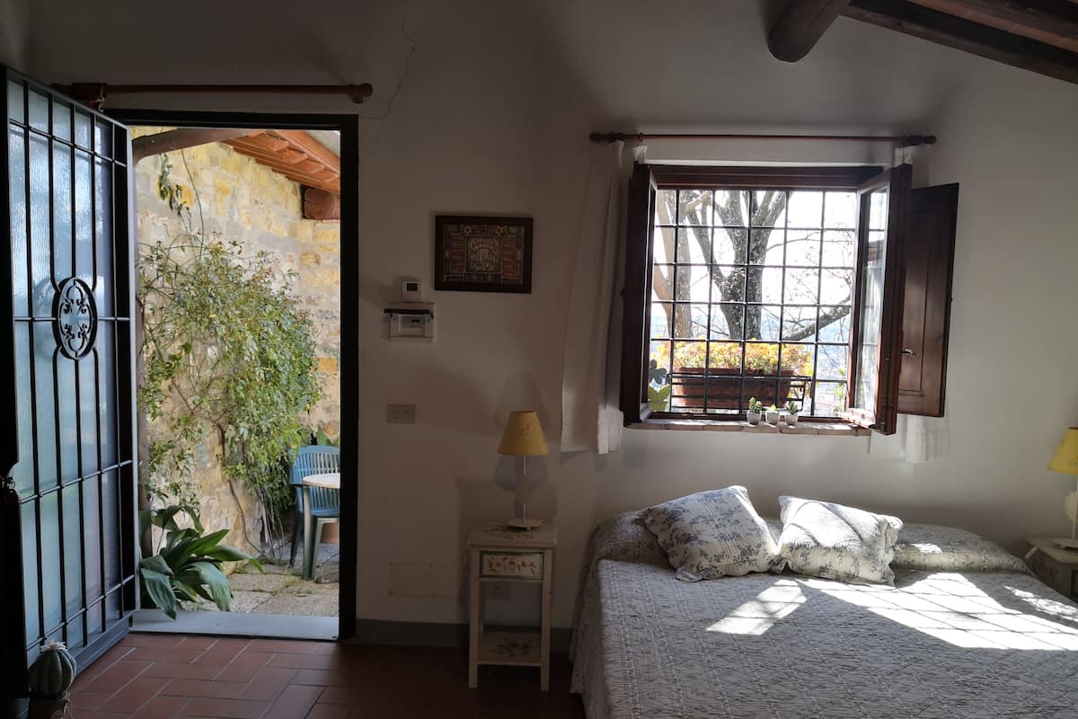 A bedroom with a bed, a window with a view of the outside, a small table with a lamp, and a door leading to a patio area.