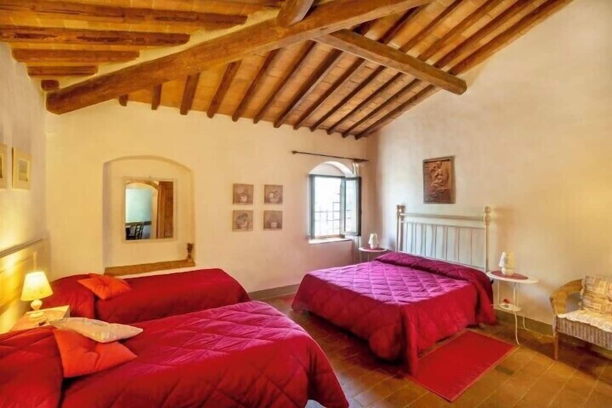 A room with two beds, wooden beams on the ceiling, a window, and a small table with a lamp.