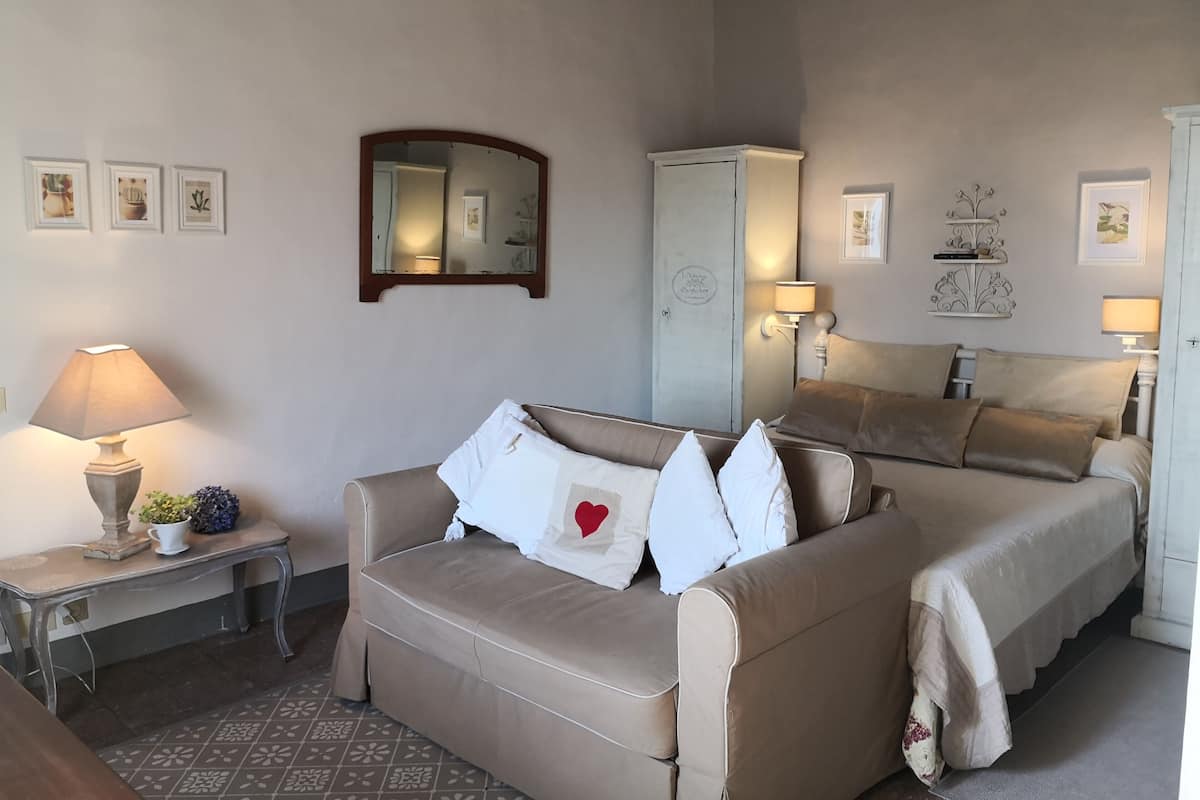 A bedroom with a beige sofa, a bed with beige bedding, a nightstand with a lamp, a mirror, and a cabinet.