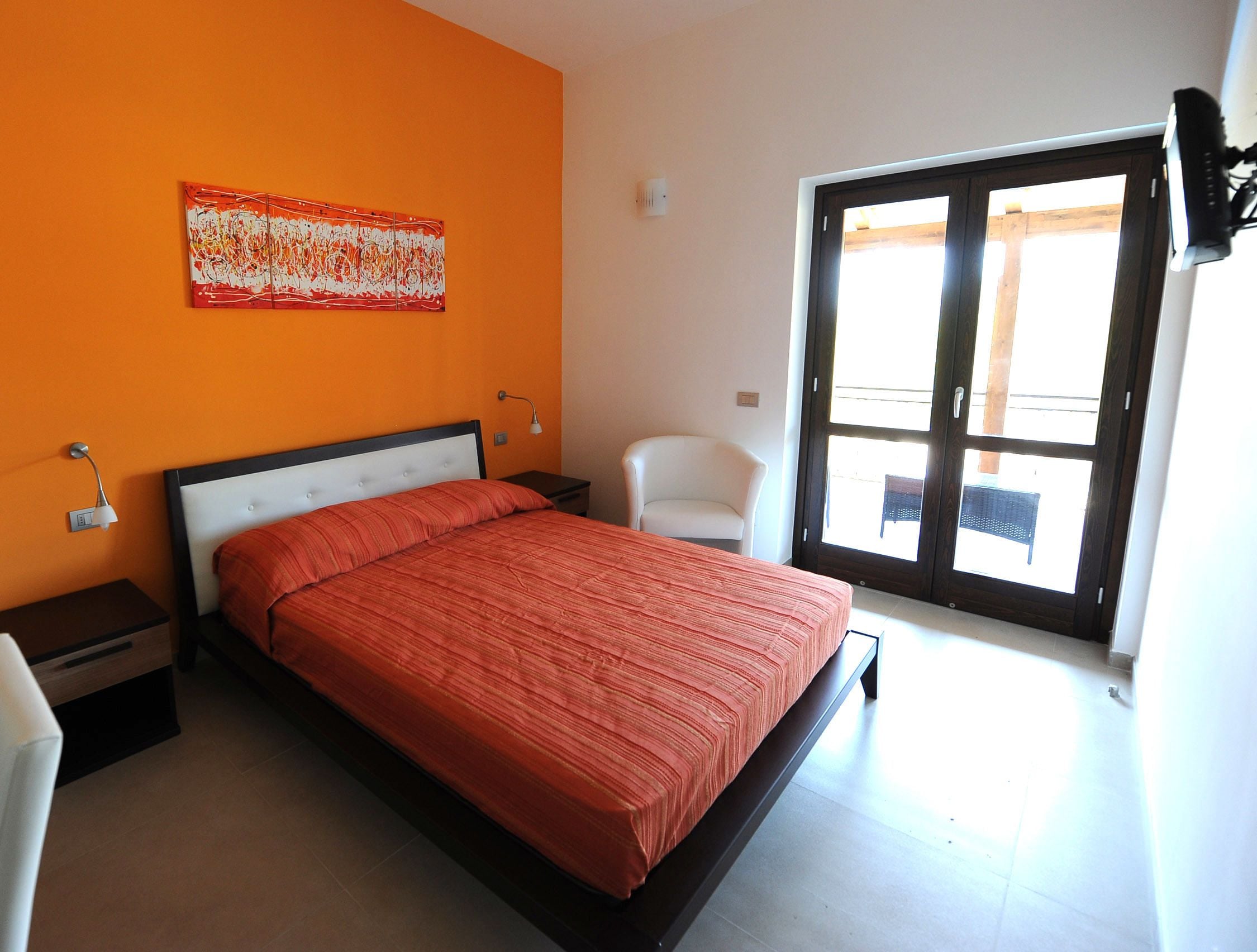 A bedroom with a bed, a chair, a nightstand, a wall art piece, and a sliding glass door.