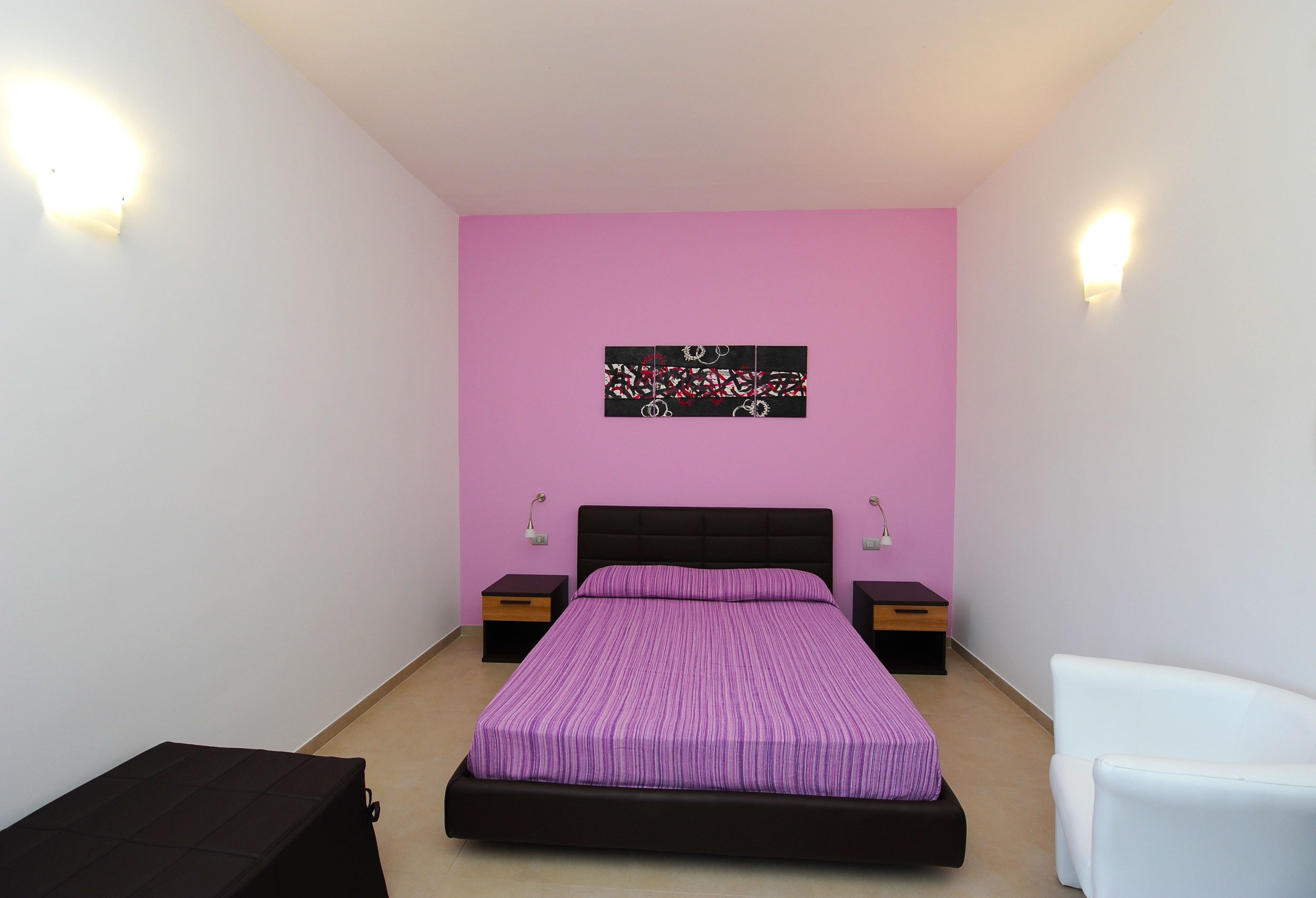 Een slaapkamer met een bed, nachtkastje, stoel en een kunstwerk aan de muur.