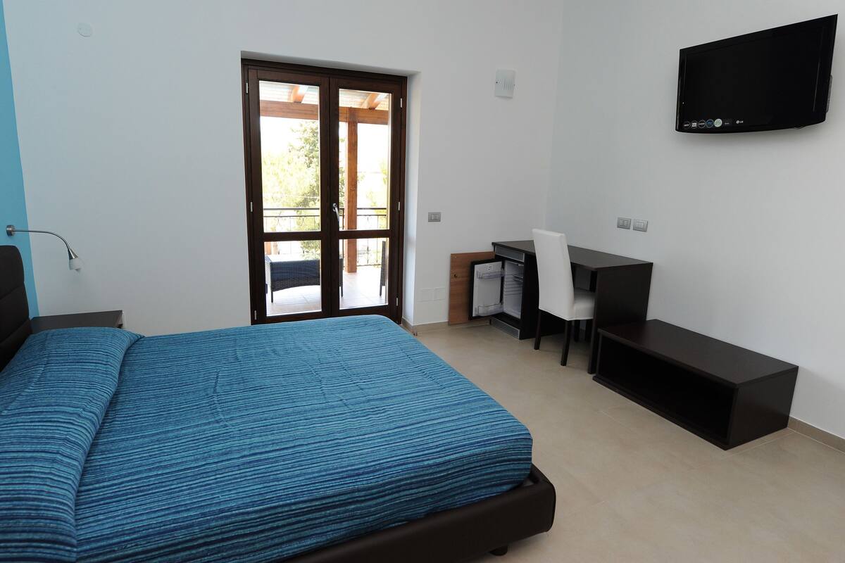 Ein Schlafzimmer mit einem Bett, einem Schreibtisch, einem Stuhl, einem Fernseher und einer Schiebetür zu einem Balkon.
