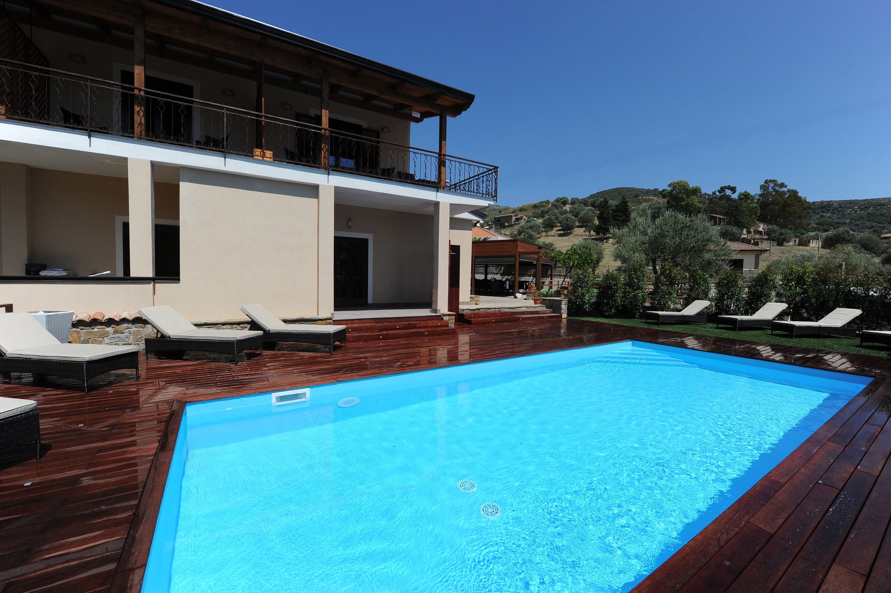 Ein Swimmingpool mit Holzterrasse und Liegestühlen, ein Gebäude mit Balkon und eine hügelige Landschaft im Hintergrund.
