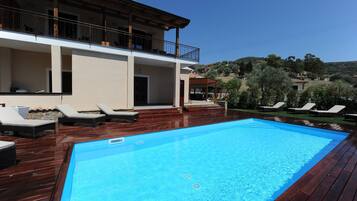 Ein Swimmingpool mit Holzterrasse und Liegestühlen, ein Gebäude mit Balkon und eine hügelige Landschaft im Hintergrund.