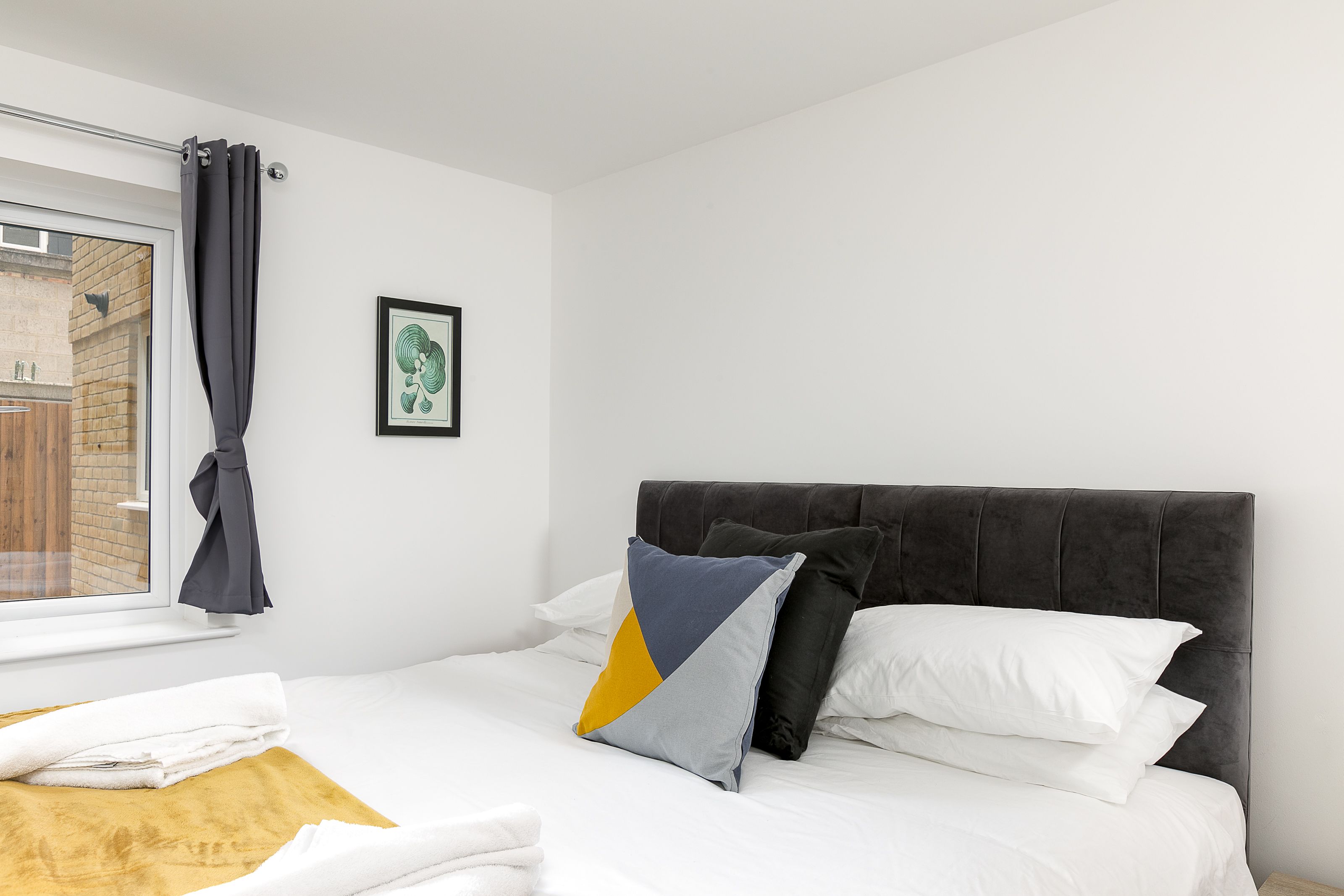 A hotel room with a bed, a window with curtains, a framed picture on the wall, and a yellow towel on the bed.
