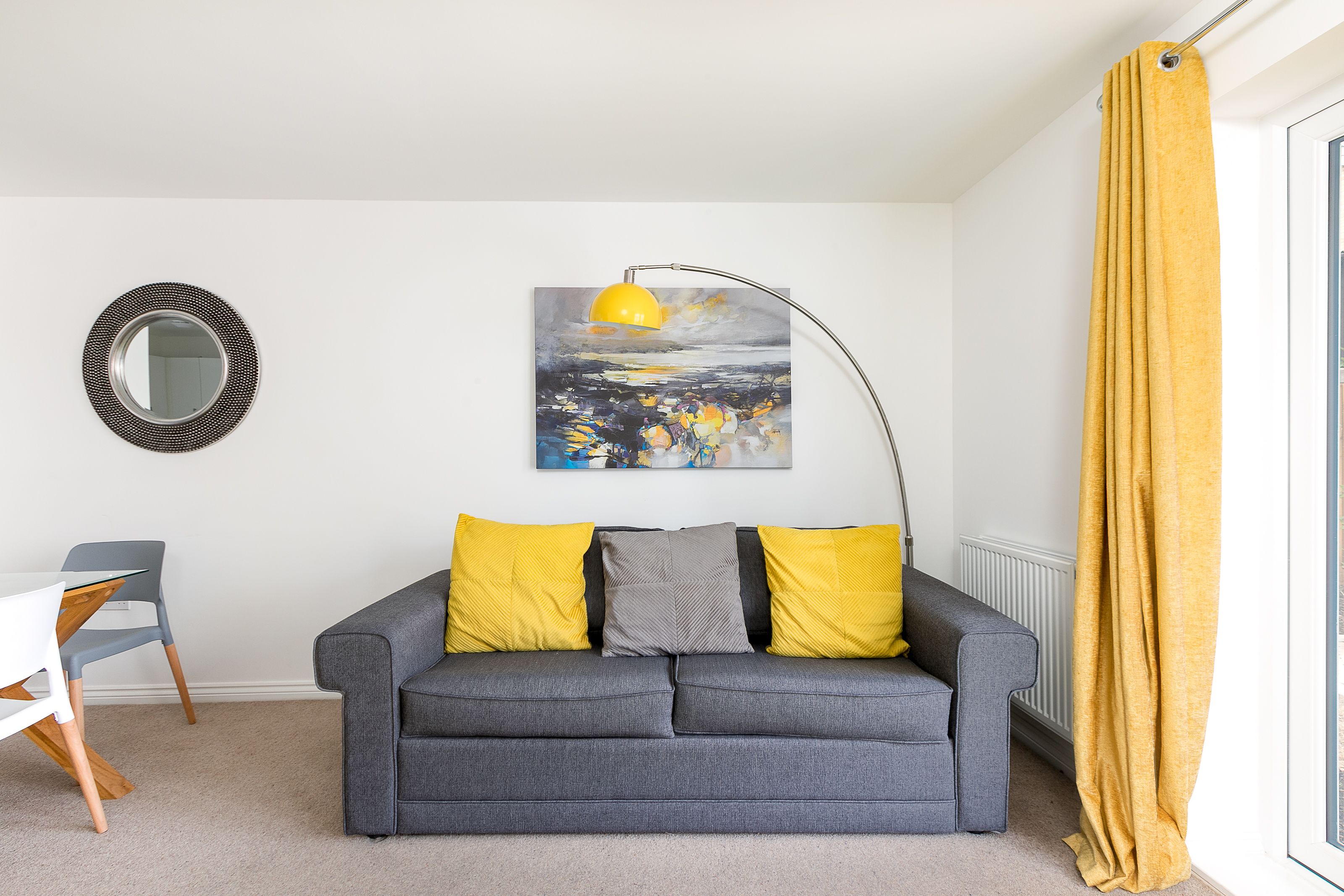 Un salon moderne avec un canapé gris, des coussins jaunes, un miroir rond et un tableau représentant un paysage.