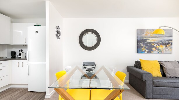A modern kitchen with white cabinets, a white refrigerator, and a dining area with a glass table and yellow chairs.