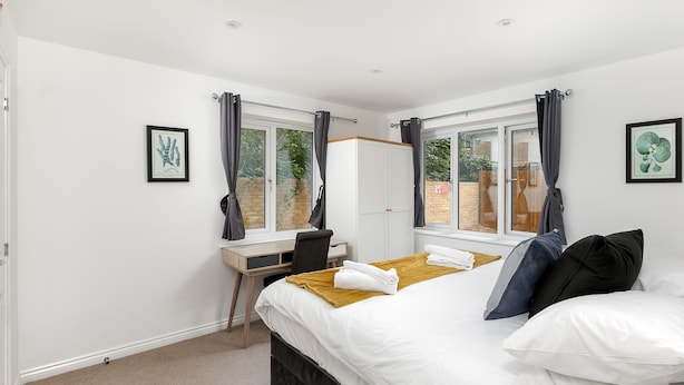 A bedroom with a bed, a desk, and two windows with curtains.