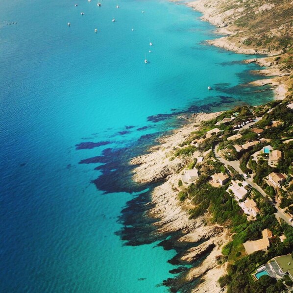An aerial view of a coastal area with clear turquoise waters, rocky terrain, and scattered buildings.