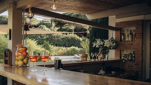 A bar area with a wooden counter, hanging light bulbs, a glass jar of fruits, and a brick wall with bottles. - La Villa d'Andrea (Ramatuelle)