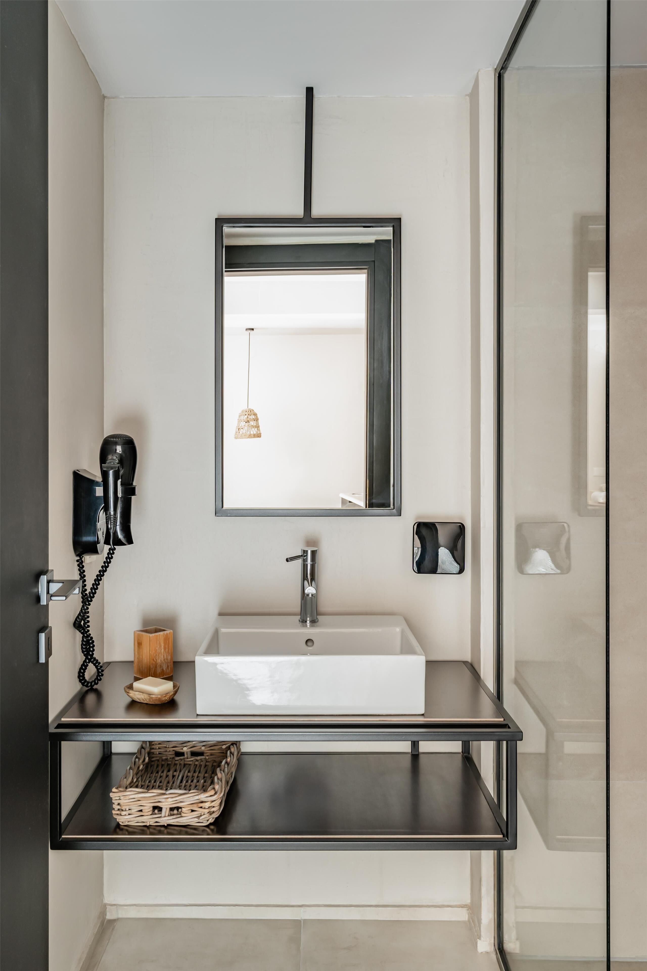 Une salle de bain moderne avec un grand miroir, un lavabo blanc, un sèche-cheveux et un panier en osier posé sur un comptoir noir.