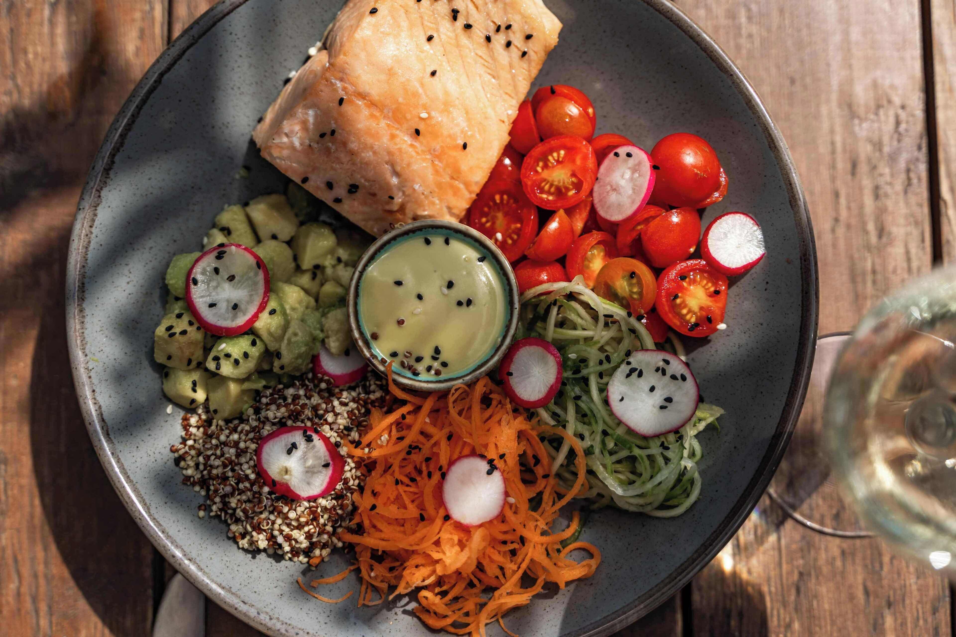 A plate with salmon, cherry tomatoes, radishes, avocado, and a bowl of sauce.
