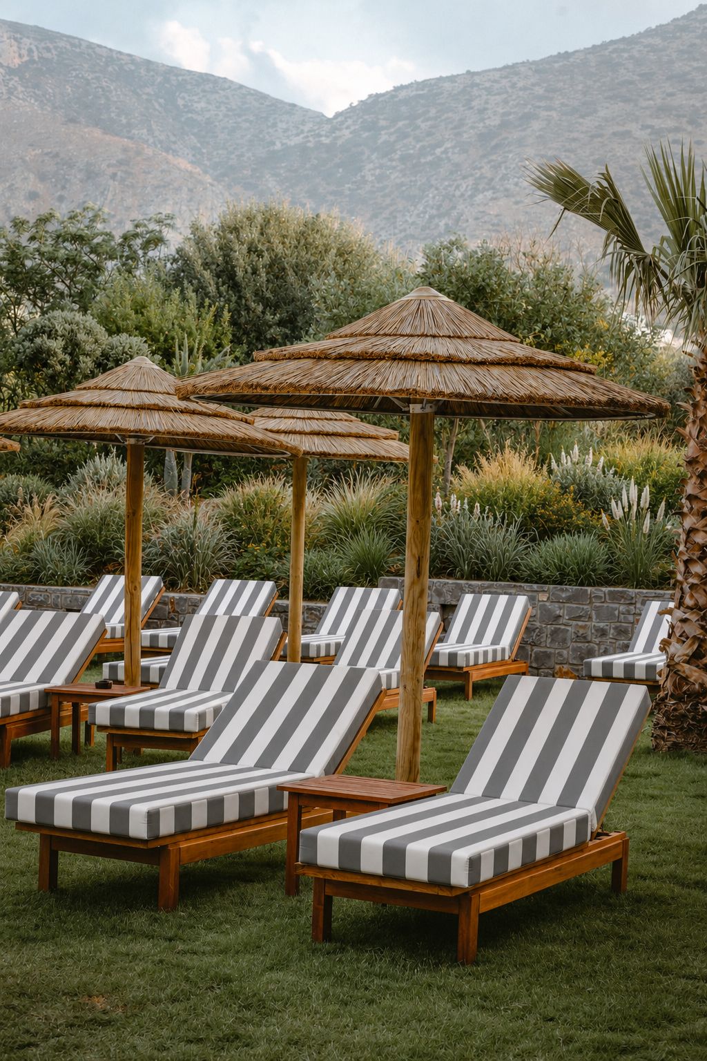 A row of striped lounge chairs with thatched umbrellas, set against a backdrop of greenery and mountains.