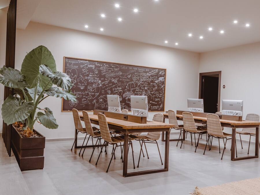 Un salón de conferencias con una gran pizarra llena de anotaciones, varias estaciones de trabajo con computadoras y una planta grande en maceta.