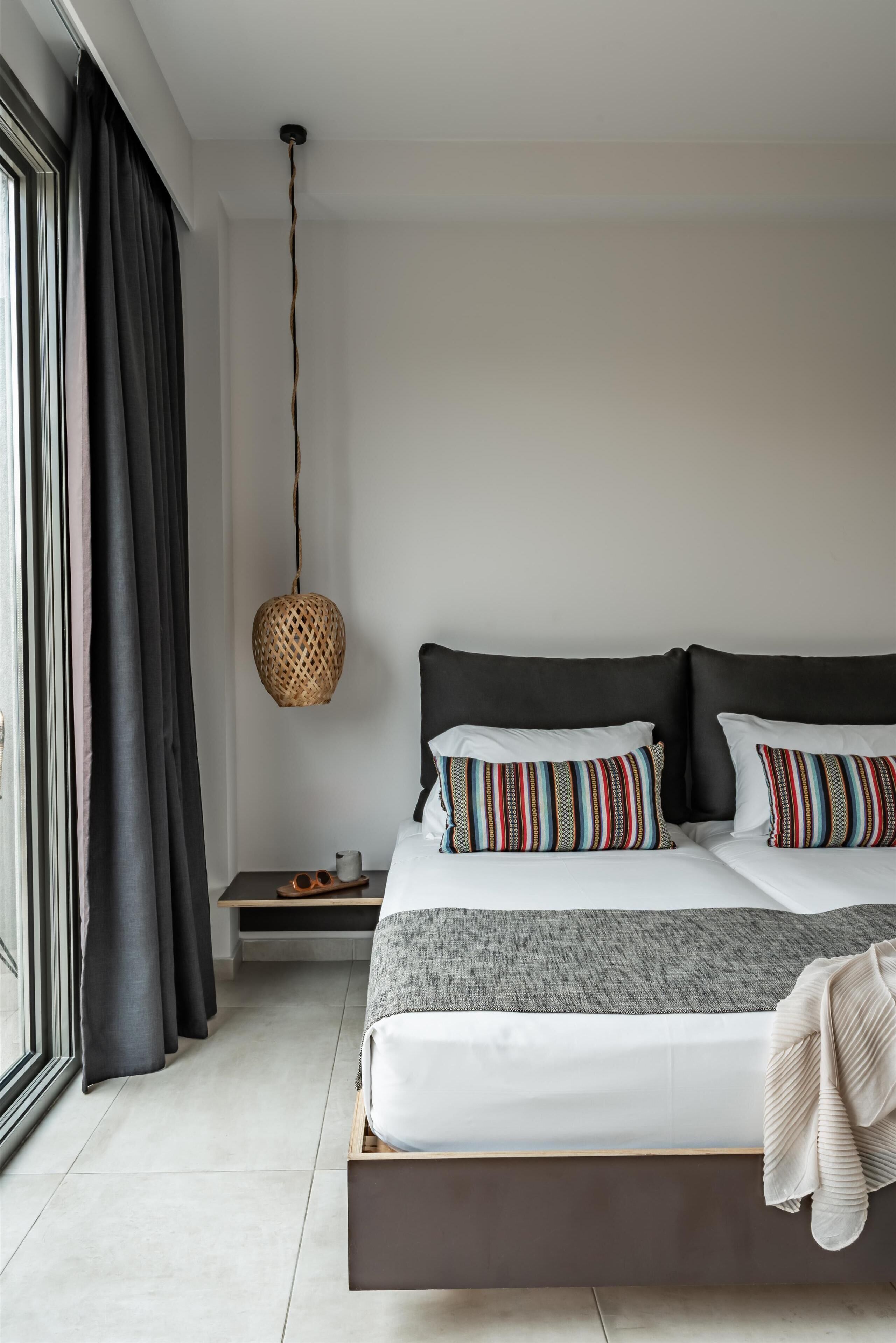 A modern bedroom with a large bed, dark curtains, a woven light fixture, and a view of the outdoors.