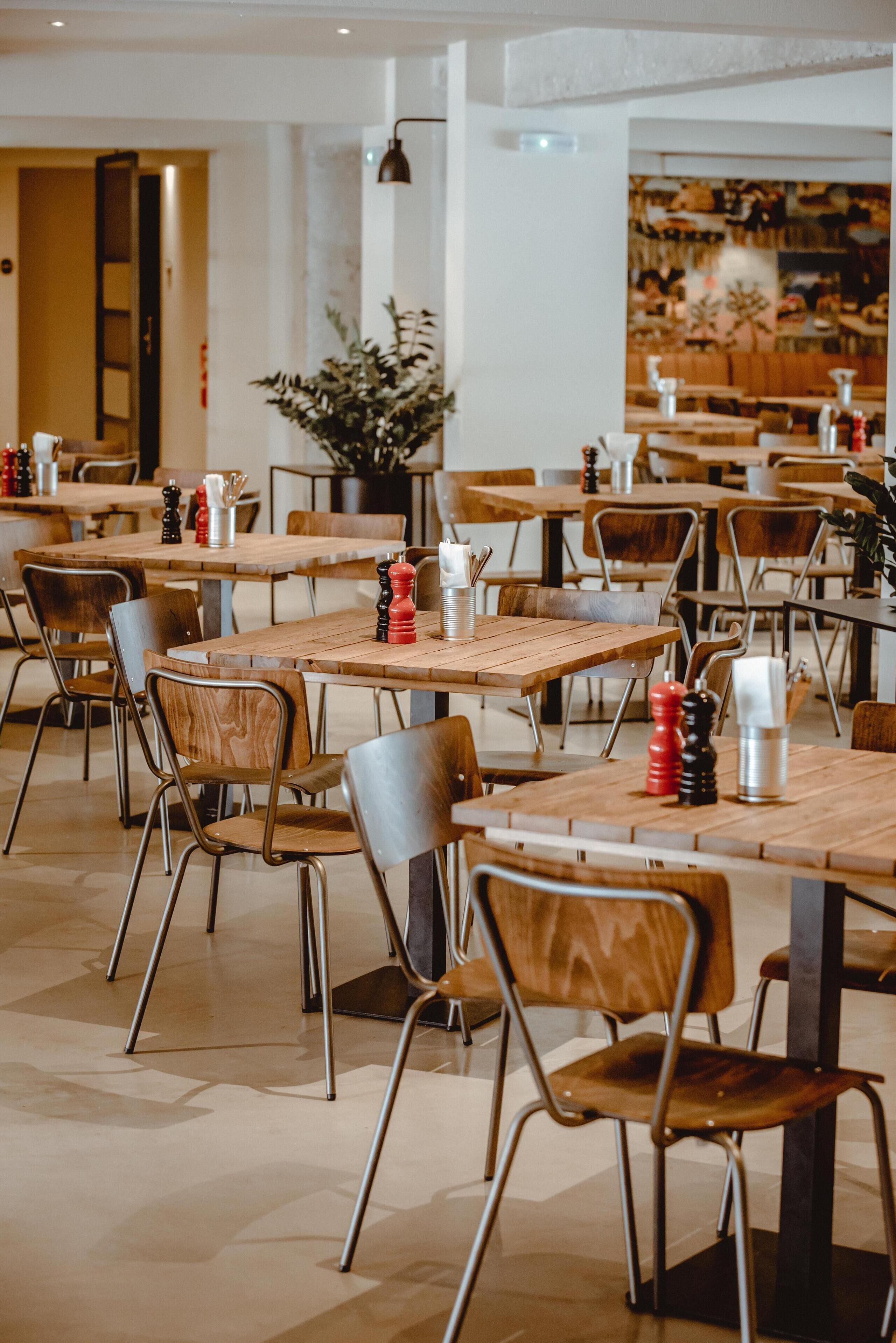 L’intérieur d’un restaurant présente de nombreuses tables et chaises en bois, une plante en pot et un mur orné d’une fresque.