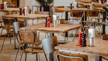 L’intérieur d’un restaurant présente de nombreuses tables et chaises en bois, une plante en pot et un mur orné d’une fresque.
