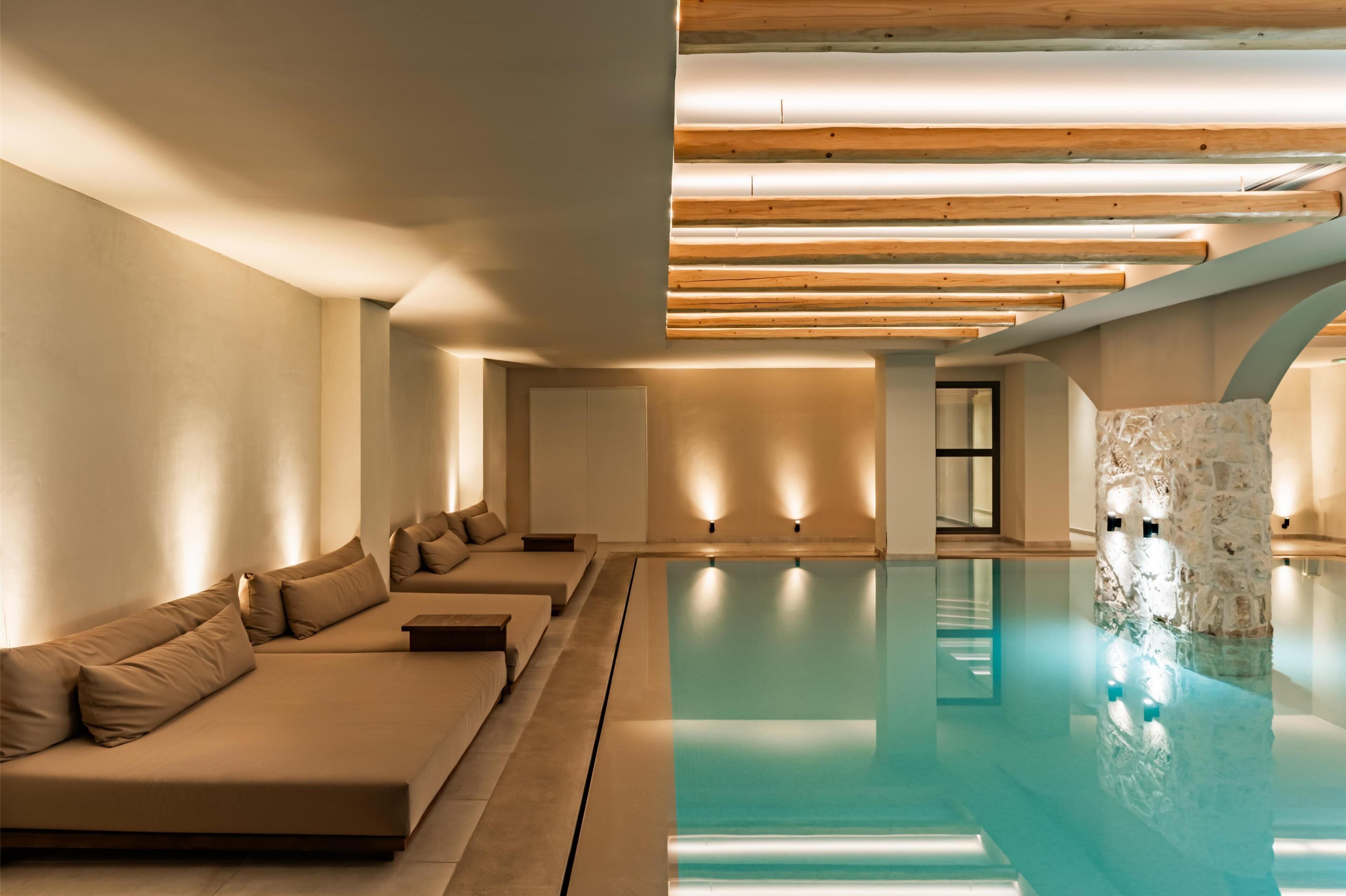 A modern indoor pool area with a long, narrow pool, a sofa with cushions, and wooden ceiling beams.