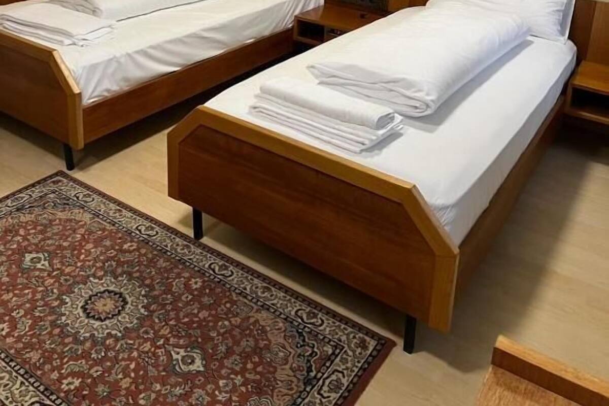 Habitación de hotel con dos camas de madera, una mesita de noche pequeña, una mesita auxiliar, un estante decorativo en la pared y una alfombra estampada.