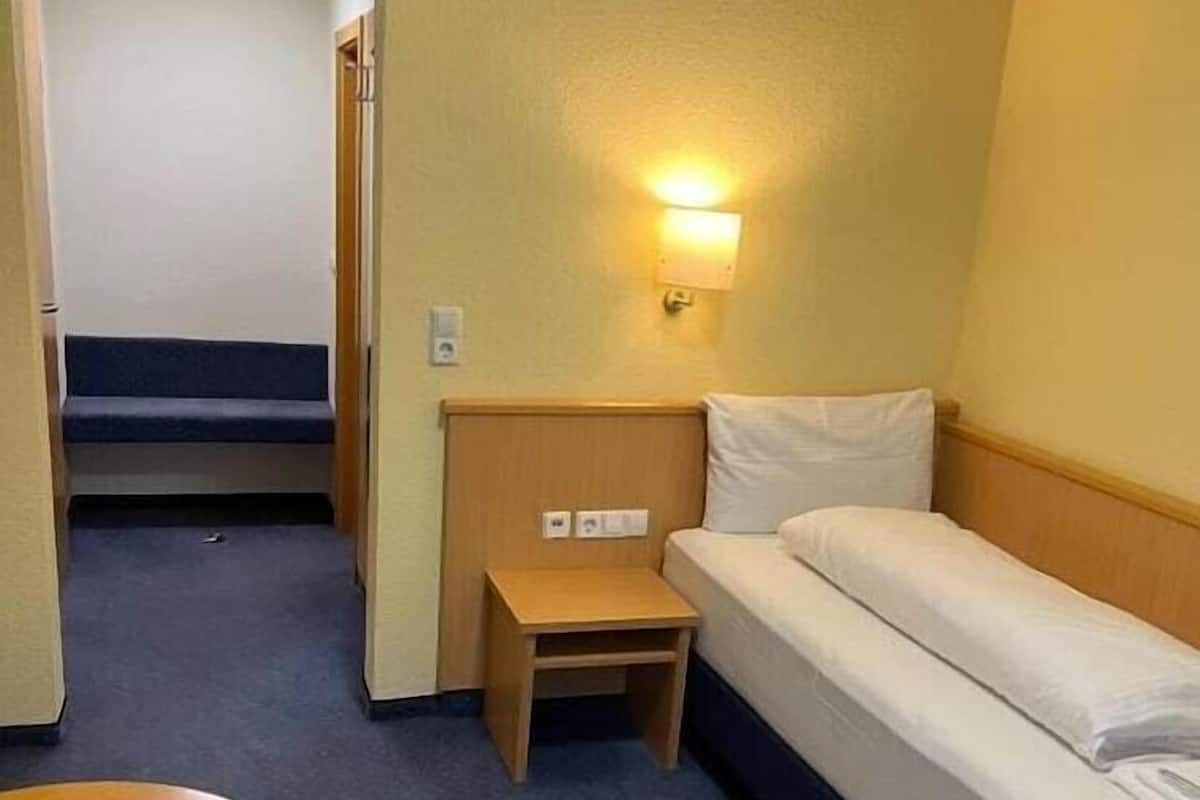 Habitación de hotel pequeña con una cama individual, una mesita, un banco y una lámpara en la pared.